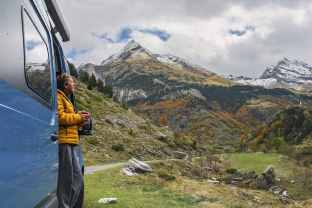 Зрелый мужчина, припаркованный в своем фургоне над Гаварни в Верхних Пиренейских горах, любуется видом на окружающие горы. Он одет в желтую куртку и держит горячий напиток. 1776564269 pic du piméné