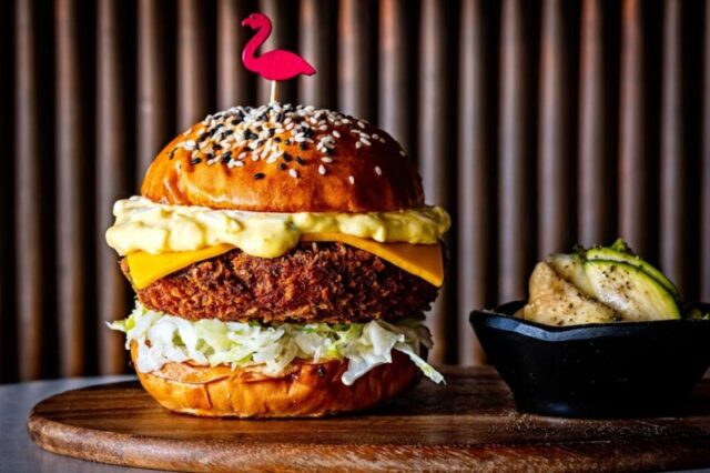 Высокий многослойный бургер с кунжутной булочкой и фламинго-скрепкой на деревянной доске рядом с маринованными огурцами в маленькой чашке. Бургер с креветками кatsu из Perry's в Вашингтоне, округ Колумбия.