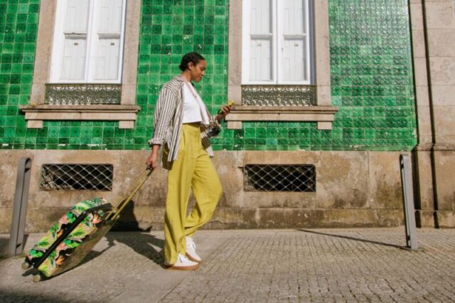 Вертикальный снимок чернокожей туристки с чемоданом, которая идет перед зеленым историческим зданием с плиткой, проверяя на своем телефоне направление в солнечный день, неся свой багаж, Тип лицензии: медиа,