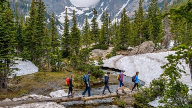 Эстес Парк, Колорадо, США - 24 июня 2017 года: В туманный весенний день группа туристов пересекает мост из дерева через ручей Тиндалл на тропе Эмеральд Лейк у подножия пика Халлетт и горы Флаттоп. 708951949